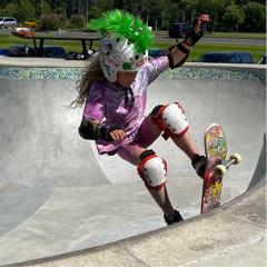 Girl in pink shirt and helmet with green mohawk skateboarding in a concrete bowl